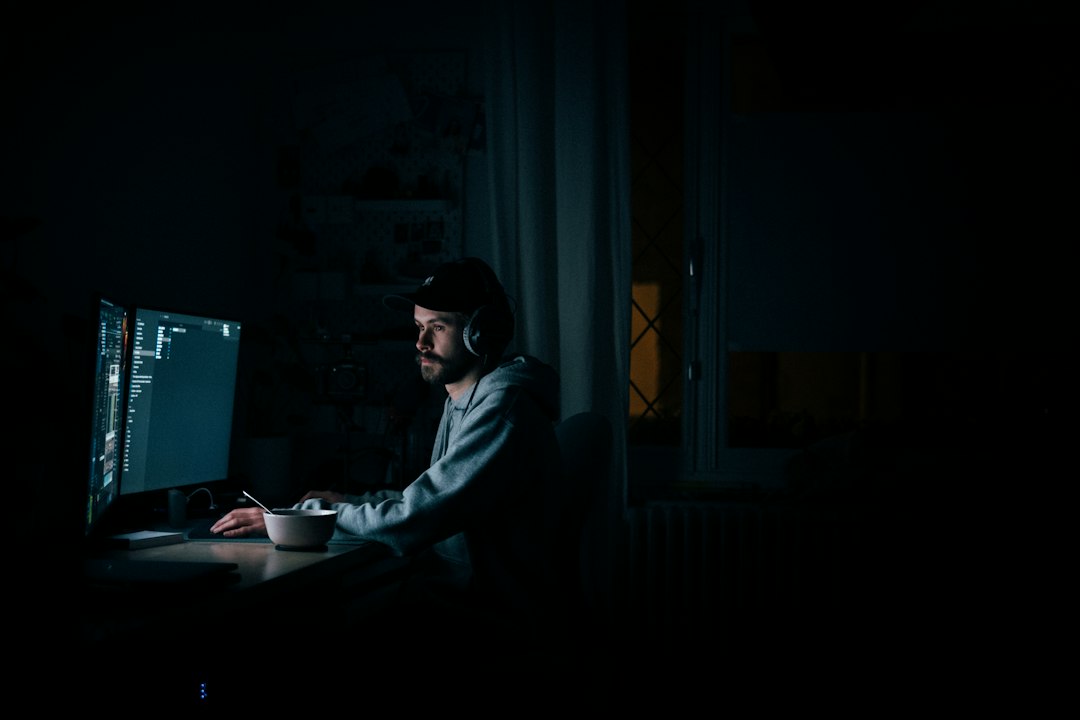 A person seen as a silhouette sitting in a dark room, their face illuminated by the chaotic light from multiple glowing computer and phone screens, representing digital information overload.